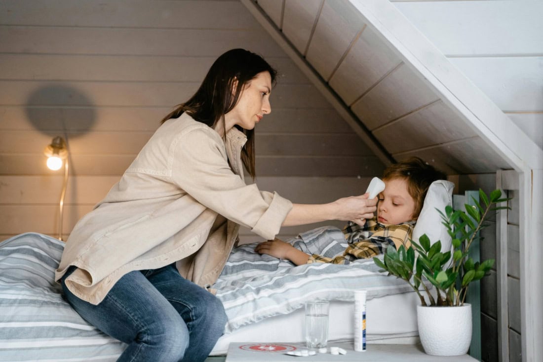 Sick child with fever is cared for by mother in bedroom, thermometer and medication present.